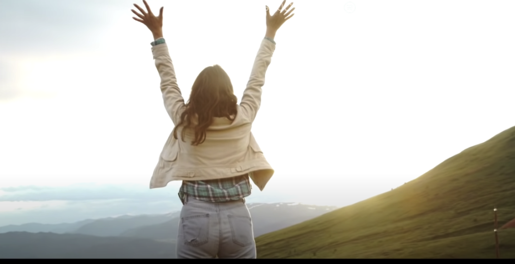 Photo of a girl raising her hand outdoors in a serene scene, surrounded by mountains and the warm sun